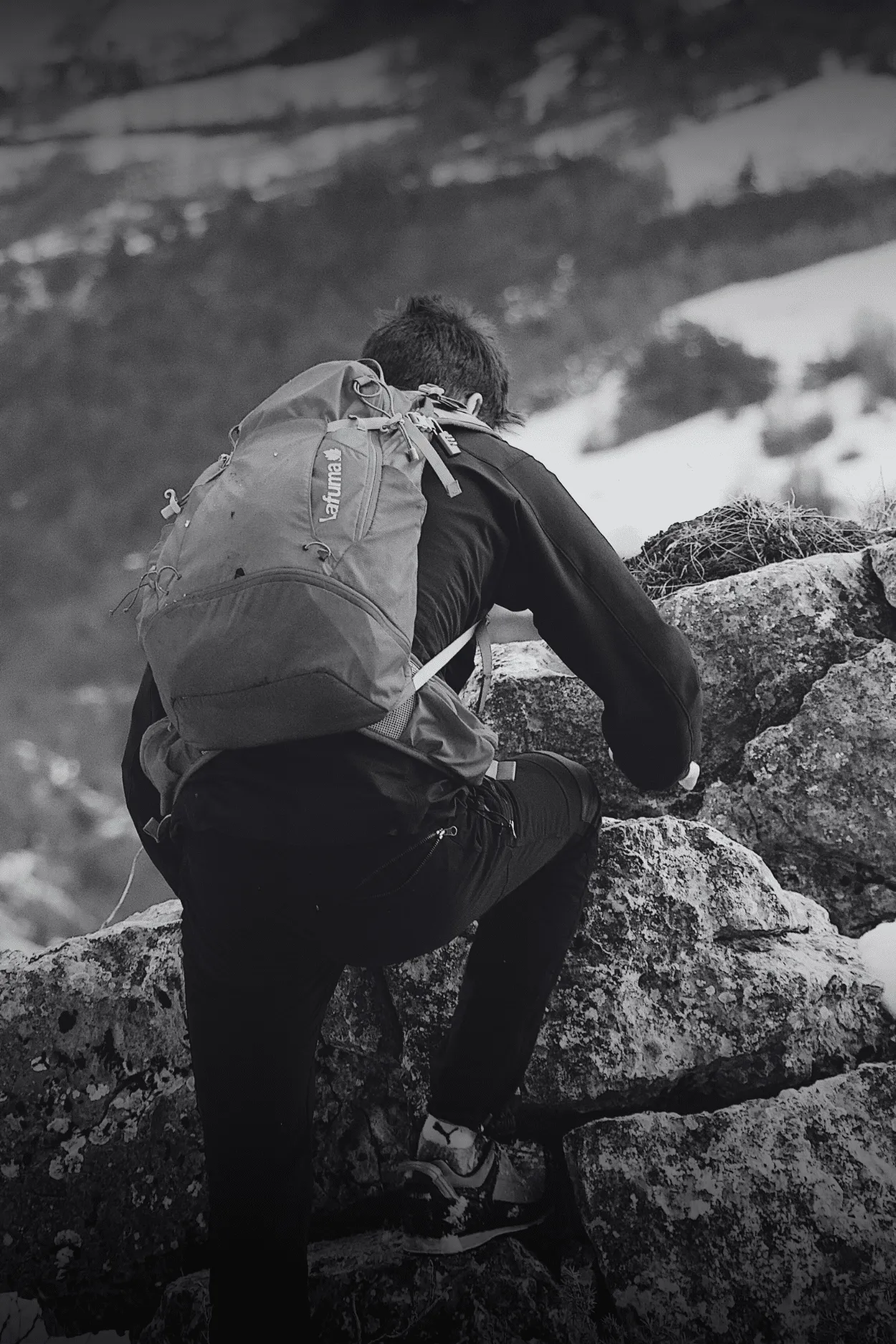 Persona con mochila trepa unas rocas, con nieve al fondo.