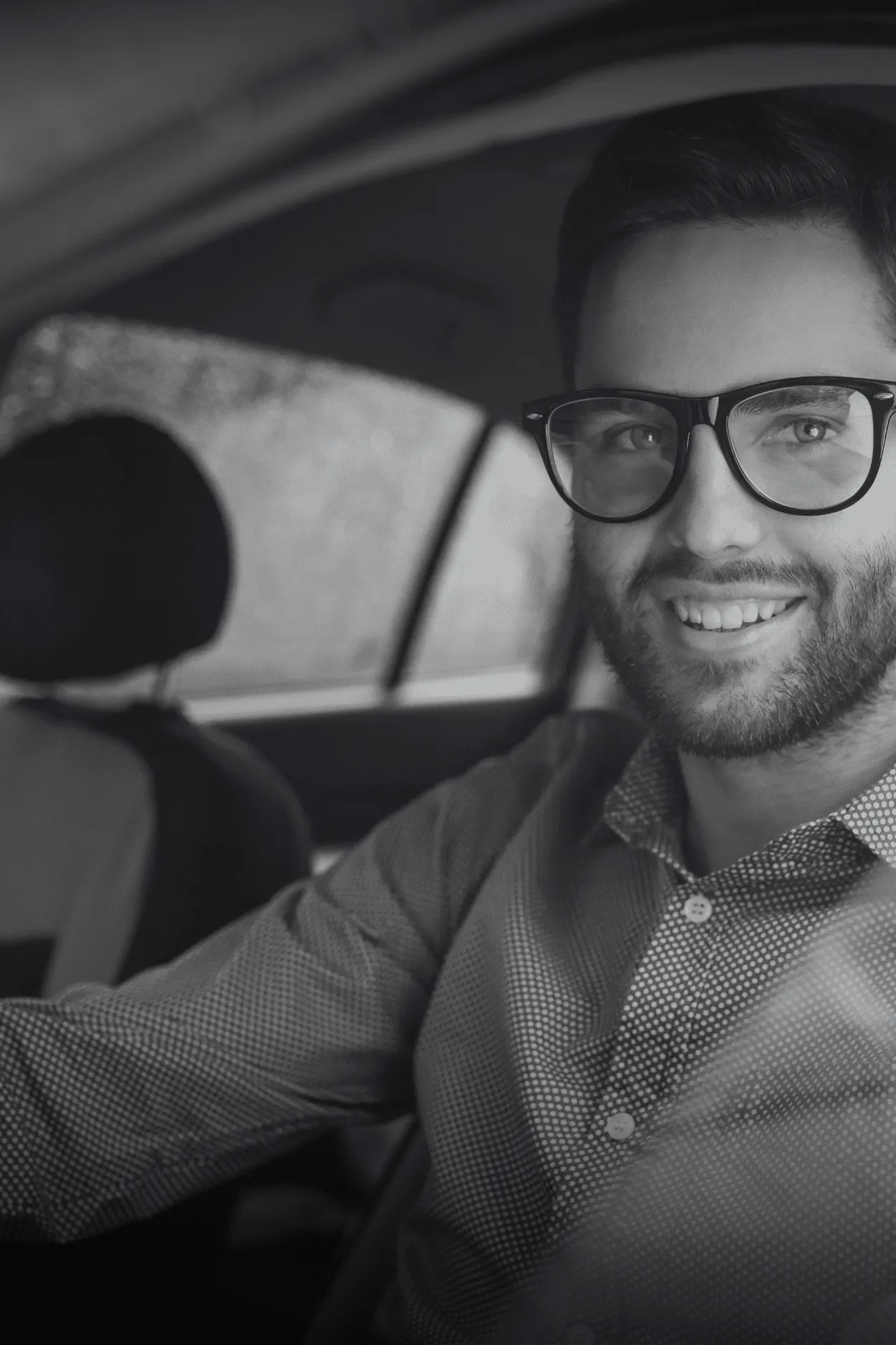 Hombre con barba y lentes sonríe al volante, con lluvia en el vidrio.
