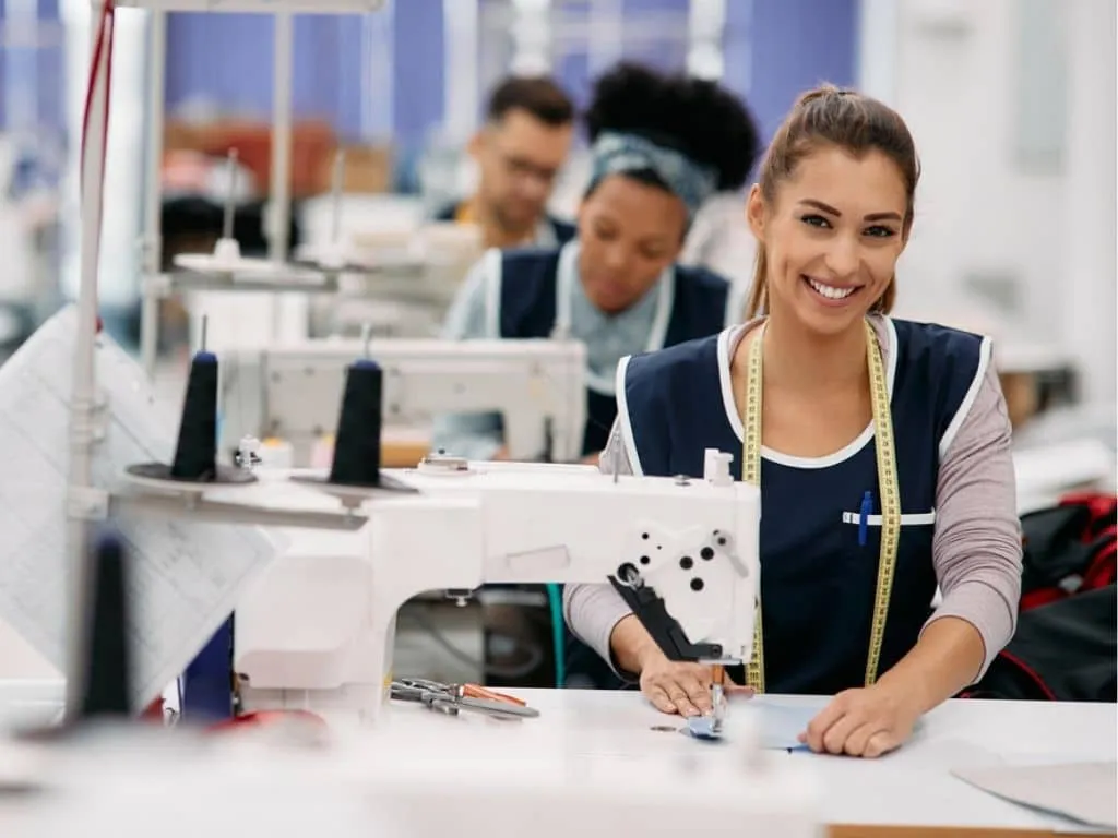 Mujer sonriente cose en una máquina industrial, con un metro al cuello y compañeros al fondo.