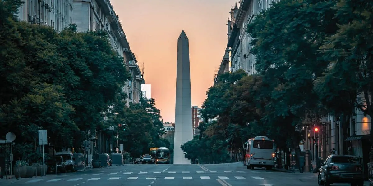 El Obelisco de Buenos Aires al fondo de una avenida, al atardecer, entre árboles y edificios.
