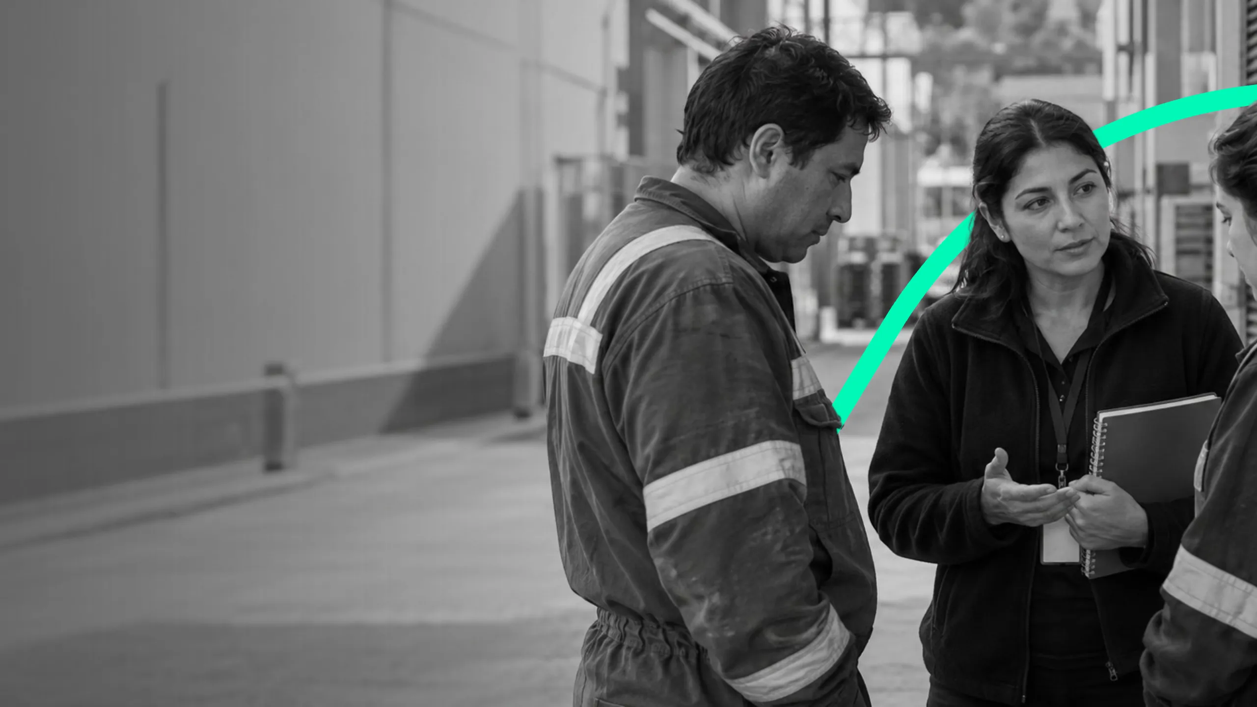 Un trabajador con overol escucha a una mujer con cuaderno en un pasillo industrial.