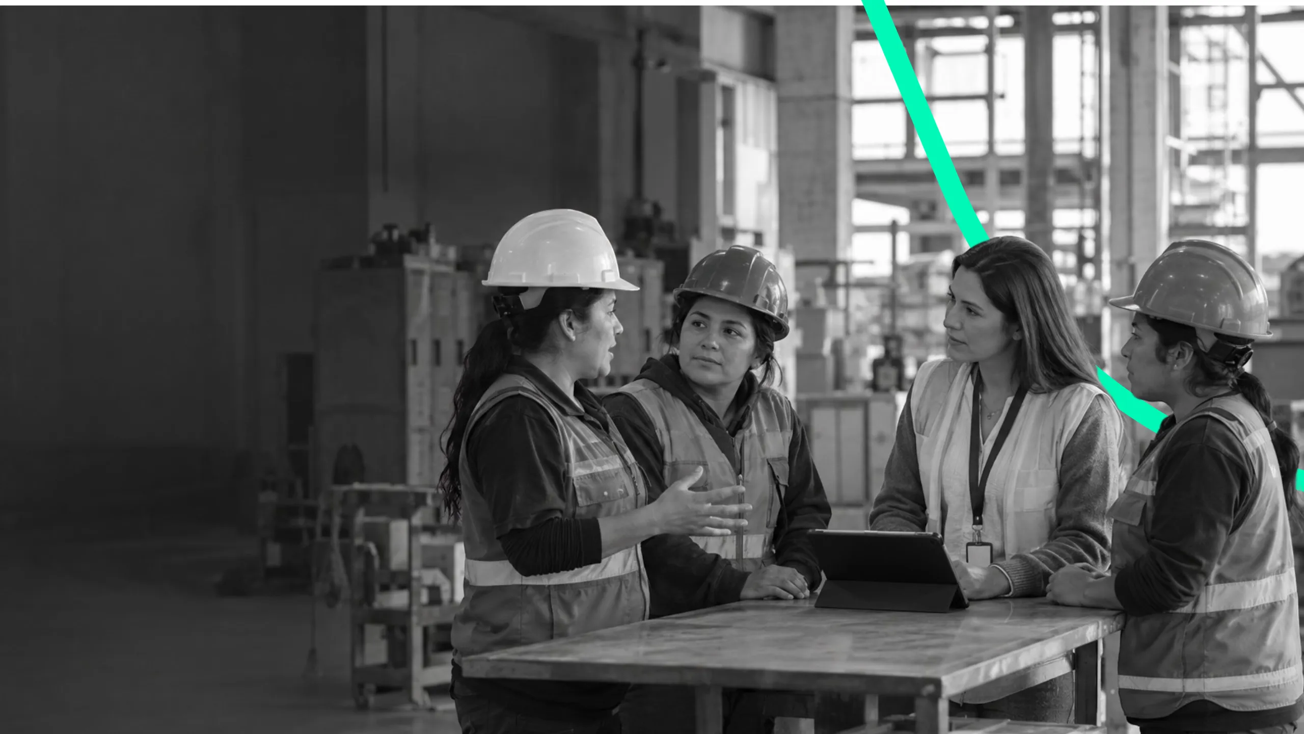 Tres trabajadoras con casco conversan con una colega sin casco en una mesa de una planta industrial.