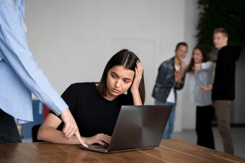 Una mujer frente a un computador se lleva la mano a la cabeza, mientras otras personas se ríen detrás.