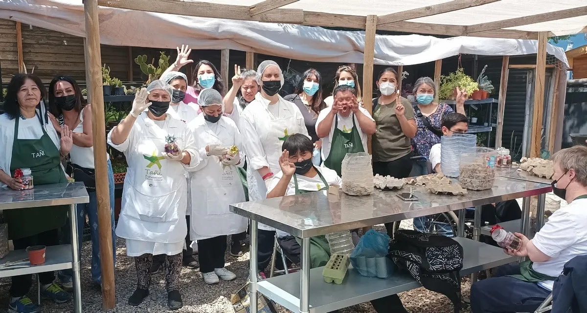 Grupo con delantal y cotona blanca saluda bajo un toldo, junto a una mesa con maples de huevo.